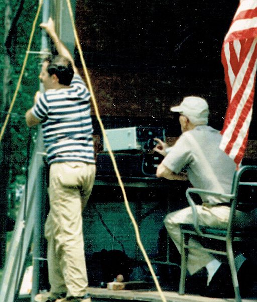 two men, one holding the mast of an antenna, the other tuning a radio (seated)